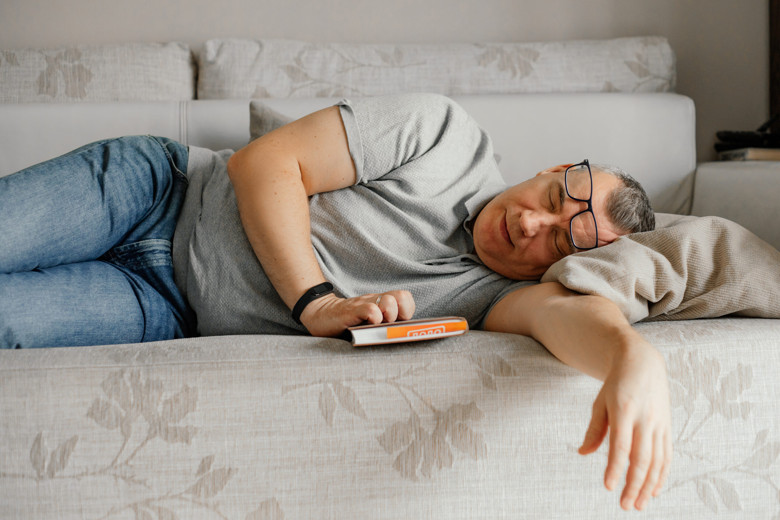 Aged man with glasses fall asleep reading book on comfortable couch in home room. Daytime sleep, napping. grandfather asleep on the couch at home