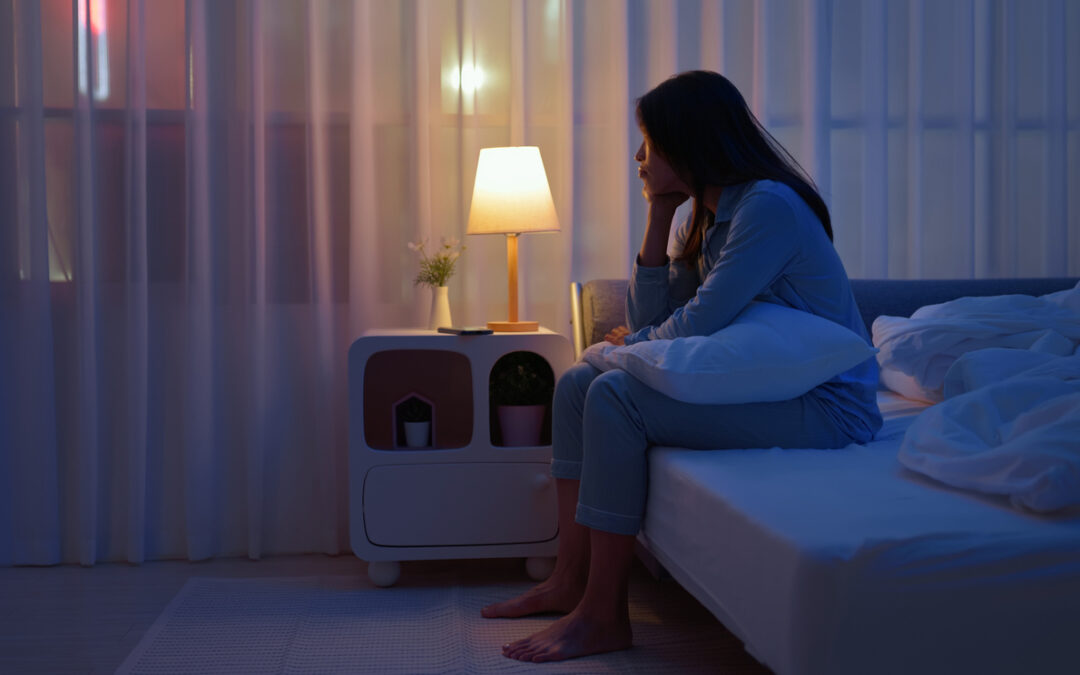 side view asian woman sits alone by the bedside in a dark room feeling troubled and unable to sleep